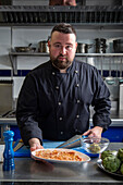 Bearded male chef in black uniform looking at camera and offering plate with burned meat while working in kitchen of contemporary restaurant
