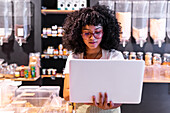Young African American woman in uniform and glasses with curly hair checking goods in glass boxes and using laptop while working in zero waste grocery store
