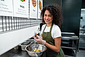 Positive ethnic female seller in gloves and apron cutting fresh vegetables while cooking salad and working in bar in supermarket