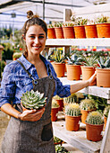 Woman in apron looking at camera smiling with checkered shirt demonstrating pot with green succulent to camera while working in greenhouse