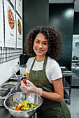 Positive ethnic female seller in gloves and apron cutting fresh vegetables while cooking salad and working in bar in supermarket