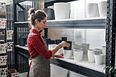 Side view of adult woman in apron arranging empty pots on shelf while working in flower store