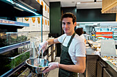 Side view of smiling male seller in apron putting green herbs in bowl while working in salad bar in supermarket and looking at camera