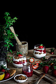 Hohe Winkel der leckeren Dessert mit Käse und frischen reifen Erdbeeren in der Zusammensetzung getopft grünen Efeu und alten Mörtel mit Stößel auf Holztisch vor schwarzem Hintergrund
