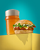 Delicious burger with fresh bun placed on yellow block near glass of cold foamy beer against turquoise background