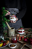 Erntehelferin in schwarzem Hemd und grauer Schürze beim Kochen eines leckeren Desserts mit Käse und frischen reifen Erdbeeren und Keksen am Tisch vor schwarzem Hintergrund