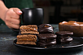 Chocolate alfajores with dulce de leche placed on black plate against blurred background of crop person holding cup