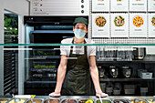 Cheerful male seller in mask and gloves standing at counter with assorted salads while working in supermarket and looking at camera