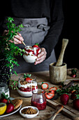 Erntehelferin in schwarzem Hemd und grauer Schürze beim Kochen eines leckeren Desserts mit Käse und frischen reifen Erdbeeren und Keksen am Tisch vor schwarzem Hintergrund