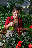 Cheerful young woman in apron spraying clean water on bright tropical flowers and smiling during work in hothouse