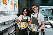 Content diverse sellers in aprons standing with freshly cooked salads in bowls while working in bar in supermarket and looking at camera