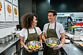 Content diverse sellers in aprons standing with freshly cooked salads in bowls while working in bar in supermarket and looking at each other