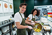 Seitenansicht von männlichen und weiblichen Kollegen in Schürzen, die in der Bar eines Supermarkts gesunde Salate zubereiten und dabei in die Kamera schauen