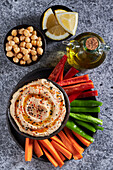 Top view of delicious hummus and slices of assorted vegetables served near bottle of oil and bowls of chickpea and lemon pieces on gray marble table