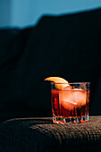 Glass of refreshing Negroni cocktail with bitter flavor and ice garnished with orange peel and served on couch arm in dark room