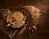 From above rustic composition of harvesting sickle on bunch of wheat ears near bowl full of golden corns on burlap on wooden table