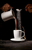 Crop unrecognizable person pouring tasty cocoa and sweet sugar into white mug placed on table in kitchen on black background