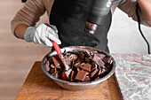 Cropped unrecognizable worker in black apron wearing gloves while melting dark chocolate in stainless steel bowl using heat gun working at counter with marble slab