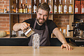Cheerful bearded male barista in apron pouring ingredient from stainless pitcher into glass with ice cubes in coffee house