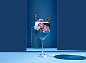 Transparent glass of fresh cocktail with mint leaves and flowers placed on surface near bottle against blue background