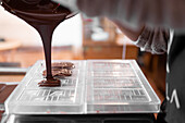 Cropped unrecognizable person in apron and gloves filling plastic mold with liquid chocolate during work in confectionery in daytime
