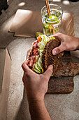 From above of crop anonymous client of restaurant eating tasty sandwich made with bacon toasts and lettuce leaves with water with lime