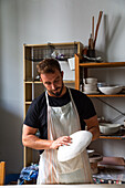 Concentrated young bearded male potter in casual clothes and apron creating white ceramic plate while working in studio