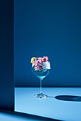 Transparentes Glas mit frischem Cocktail, Minzblättern und Blumen vor blauem Hintergrund auf dem Tisch