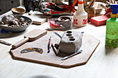 Von oben ein Stück Ton auf einem Holzbrett mit verschiedenen Bildhauerwerkzeugen und einer Metalltöpferrippe in einer Werkstatt