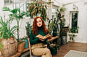 Concentrated red haired female sitting on chair with crossed legs and reading book among different potted plants in domestic garden
