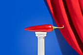 Red hot pepper placed on miniature column against blue background with red velvet curtain