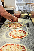 Nicht erkennbarer männlicher Koch, der bei der Arbeit in einer Restaurantküche geriebenen Käse auf einen Pizzaboden mit Tomatensauce streut