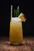 Wooden table with glass of yellow cocktail with ice cubes and refreshing cocktail garnished with spiky leaves and striped straw