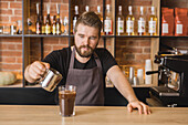 Serious bearded male barista in apron adding ingredient into glass with iced beverage in coffee house during working day