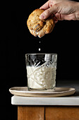 Crop anonymous person dipping fresh sweet cookie into glass of milk placed on corner of table against black background in kitchen