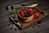 From above bowl with berries near uncooked oatmeal served on wooden cutting board with various fresh berries and mint leaves in kitchen