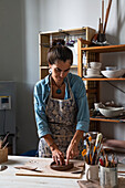 Concentrated middle aged ethnic female in casual clothes and apron shaping clay piece placed on wooden board near various pottery tools while standing at table in workshop