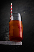 Transparentes Glas mit rotem Alkohol-Cocktail, serviert mit gestreiftem Strohhalm, an einer Ecke eines Marmortisches vor grauem Hintergrund im Studio