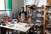 Positive middle aged female and young male in casual clothes and aprons smiling while creating clay plate during pottery class in workshop