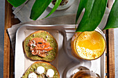 From above of delicious avocado toasts with salmon and burrata cheese served on table with glasses of fresh juice and herbal tea placed near exotic Monstera deliciosa plant