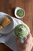 From above unrecognizable person holding glass of fresh dalgona coffee with matcha and Japanese tea ceremony accessories consisting of bamboo whisk called chasen and traditional scoop named chashaku placed on wooden table