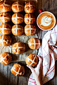 From above hand with appetizing freshly baked sourdough hot cross buns and mug of coffee on wooden table
