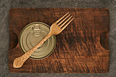 From above scratched chopping board with fork and sealed can with preserved food on rustic lumber table