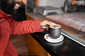 Unrecognizable Hispanic male smiling and taking cup of traditional champurrado drink from saucer with cocoa beans while sitting at counter in cafe