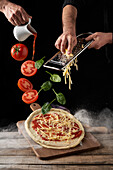 Koch reibt Käse und gießt Marinara-Sauce, während Tomaten und Basilikumblätter auf rohe Pizza vor schwarzem Hintergrund fallen