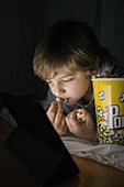 Little boy lying down comfortable on sofa with popcorn bucket while eating and watching cartoon on tablet