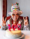 Vergnügte Seniorin mit lustigem Hut und Sonnenbrille, die mit beiden Händen das V-Zeichen zeigt und ihren Geburtstag mit einem Kuchen mit 100 Kerzen feiert