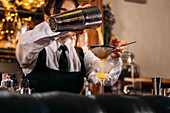 Skilled young female bartender using strainer and pouring orange alcohol drink from shaker into glass while preparing refreshing cocktail at bar counter