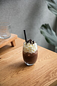 Glas mit Schokoladenfrappé mit süßer Schlagsahne, serviert mit Strohhalm auf einem Holztisch in einem hellen Raum