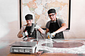 Hispanic woman in black apron and cap melting and stirring chocolate in bowl near male colleague spreading substance on board and measuring temperature with infrared thermometer during work in confectionery